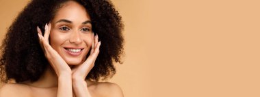 Mutlu genç siyahi bayanın yanaklarına dokunduğu, lüks organik güzellik ürünleri, bej arka plan, yakın plan, kopyalama alanı kullandıktan sonra ipeksi teninin tadını çıkardığı vesikalık fotoğraf.