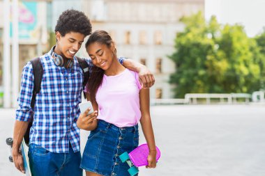 Bu görüntü genç siyah bir çiftin bir şehir caddesinde yan yana durduğunu gösteriyor. Akıllı telefona bakıyorlar ve gülüyor gibi görünüyorlar, uzayı kopyalayarak.