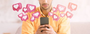 Bir adam kanepede oturuyor, elinde bir akıllı telefon tutuyor, ekrana bakıyor. Sosyal medya tebligatlarını hatırlatan bir dizi pembe kalp simgesi etrafında uçuşuyor..
