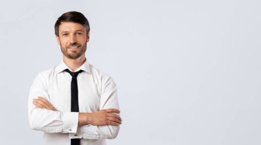 Beyaz gömlekli ve siyah kravatlı bir adam kollarını beyaz bir arka plana kavuşturmuş duruyor. Kameraya güvenle gülümsüyor..