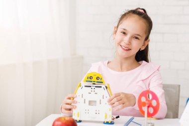 Sarı, mavi ve beyaz plastikten yapılmış robot bir oyuncak tutarken at kuyruklu genç bir kız gülümsüyor. O, önünde diğer inşaat malzemeleriyle birlikte bir masada oturuyor..