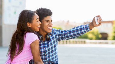 Genç siyah bir çift güneşli bir günde kentte selfie çekiyor. Genç adam telefonu tutuyor ve fotoğrafı çekerken gülümsüyor. Genç kız ona yakın duruyor.