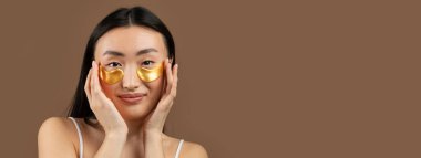Mutlu Koreli kadın göz bandı takıyor, kahverengi arka planda prosedürlerin tadını çıkarıyor, stüdyo çekimi. Cilt bakımı, doğal güzellik, kaplıca tedavisi konsepti, kopyalama alanı