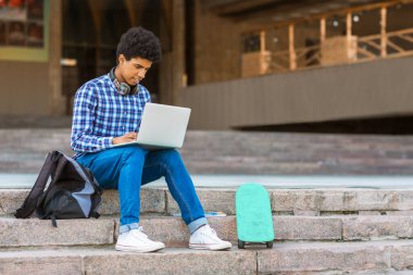 Kısa, kıvırcık saçlı genç bir zenci binanın önündeki beton basamaklarda dizüstü bilgisayar kullanarak oturuyor. Yanında siyah bir sırt çantası oturuyor ve basamakta bir deniz mavisi kaykayı duruyor.
