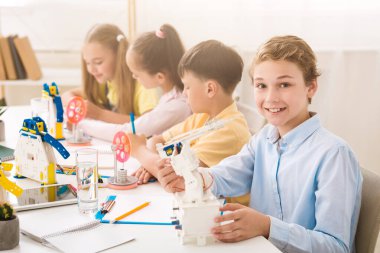 A young boy smiles at the camera while holding a robot during a STEM class. He is sitting at a table with other children who are also working on STEM projects. The classroom is bright and airy clipart