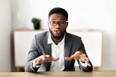 Takım elbiseli bir adam masaya oturur, elleriyle işaret eder ve doğrudan kameraya bakar..