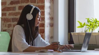 Rahat bir e-öğrenme. Kahve içen Asyalı öğrenci kafede dizüstü bilgisayarda yazı yazıyor.