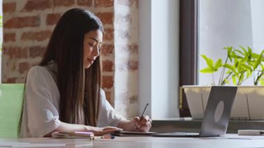 Online eğitim. Bilgisayardan Webinar izleyen ve not alan Asyalı kız kampüs kütüphanesinde oturuyor.