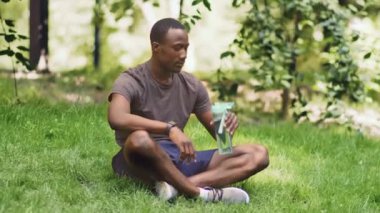 Su dengesi kavramı. Genç Afrikalı Amerikalı adam spor şişesinden içiyor, parkta çimlerin üzerinde dinleniyor.
