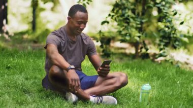 Park yerinde dinlen. Milenyum Afrikalı Amerikalı adam akıllı telefondan bakıyor, çimlerin üzerinde oturuyor.