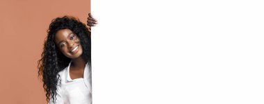 Uzun kıvırcık siyah saçlı, kameraya bakarken gülümseyen genç Afrikalı Amerikalı kadın. Büyük beyaz bir panelin arkasından bakıyor ve beyaz düğmeli bir gömlek giyiyor.