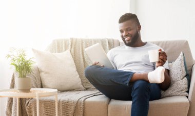 Siyah adam bir elinde tablet, diğer elinde kahve fincanı tutarken bacakları havada bir kanepede oturuyor, gülümsüyor. Kanepe ışıl ışıl aydınlatılmış bir oturma odasında.