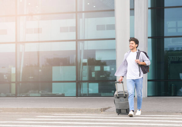 Ручной молодой человек выходит с сумкой из аэропорта, несет чемодан и переходит дорогу к автобусной остановке, копирует пространство
