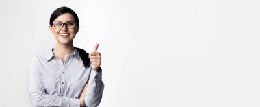 Açık mavi yakalı genç bir kadın gülümsüyor ve beyaz bir arka plana başparmak işareti yapıyor..