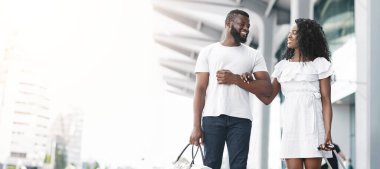 Siyah çift güneşli bir günde modern bir binanın dışında yürür. Adam büyük bir el çantası taşıyor. Kadın fırfırlı beyaz bir elbise giyiyor ve elinde bir bavul tutuyor.