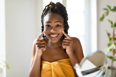 Siyah örgülü bir kadın yüzüne krem sürerken aynaya gülümsüyor. Kadın sarı bir havlu giyiyor ve parlak, beyaz bir banyoda duruyor..