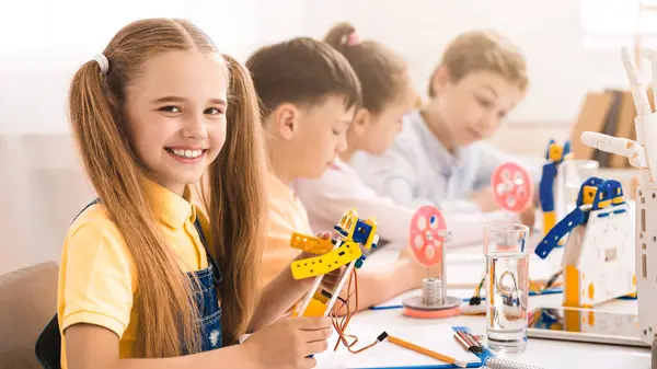 stock image A young girl with blonde hair tied up in pigtails smiles as she builds a robot during a classroom activity. She is surrounded by other children who are also working on their own projects.