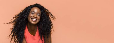 Uzun, kıvırcık saçlı, gülümseyen siyah bir kadın şeftali rengi arka planda mercan kolsuz bir bluz giyiyor. Saçları rüzgarda uçuşuyor ve kameraya doğru bakıyor.