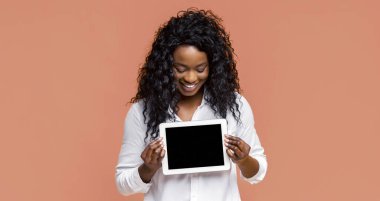 Siyah kıvırcık saçlı genç bir kadın, şeftali arkaplanının önünde siyah ekranlı beyaz bir tablet bilgisayarı tutarken parlak bir şekilde gülümsüyor.