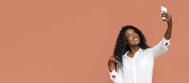 Uzun, kıvırcık siyah saçlı genç siyah bir kadın şeftali renkli bir duvarın önünde akıllı telefonuyla selfie çekiyor. O beyaz düğmeli bir gömlek giyiyor ve parlak bir şekilde gülümsüyor..