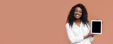 Uzun kıvırcık saçlı genç siyah bir kadın şeftali renkli bir arka planın önünde beyaz bir tablet tutarken gülümsüyor. Tablet ekranı siyah ve kadın beyaz düğmeli gömlek giyiyor..