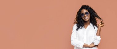 Uzun kıvırcık siyah saçlı, şeftali rengi bir arka planın önünde dururken güneş gözlüğü takan siyah bir kadın. Kadın gülümsüyor ve elinde bir tutam saç tutuyor..