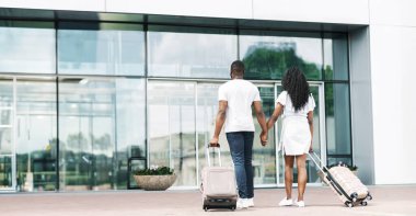 Afro-Amerikalı bir çift havaalanının girişinden çıkarken, her biri arkalarında tekerlekli bavul taşıyor. El ele tutuşup kameradan uzaklaşıyorlar.