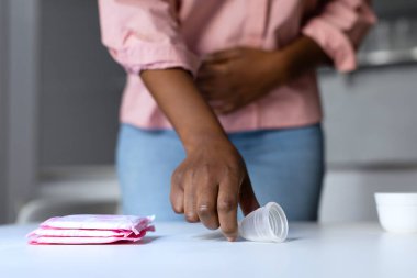 Pembe tişörtlü ve kot pantolonlu Afro-Amerikan kadın mutfağında duruyor ve bir bardak menstrual almak için uzanıyor..