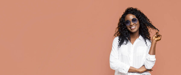Черная женщина с длинными кудрявыми черными волосами в солнечных очках, стоя на фоне персикового фона. Женщина улыбается и держит прядь волос в руке.