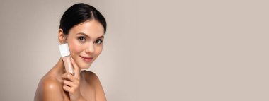 Güzel, yarı çıplak, elinde güzellik ürünü olan genç bir kadın renkli stüdyo arka planına izole edilmiş makyaj yapıyor.