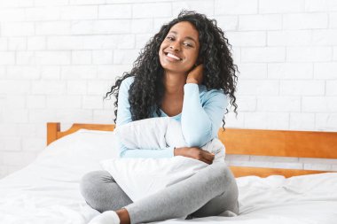 Genç Afrikalı neşeli genç bir kadının hayat tarzı portresi. Yatakta oturuyor ve gülümsüyor, yastığa sarılıyor.