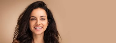 Bej renkli stüdyo arka planında poz veren güzel, uzun saçlı genç bir kadının portresi. Kameraya gülümsüyor.