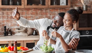Siyah adam ve kızı mutfakta cep telefonu ile selfie alarak, akşam yemeği yaparken iyi vakit geçirmek, boşluk kopyalamak
