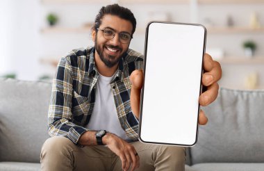 Cheerful Indian Guy Showing Smartphone With Big Blank White Screen At Camera, Happy Young Eastern Man Recommending New Mobile App Or Website While Sitting On Couch At Home, Creative Collage, Mockup