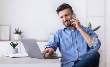 Cep telefonu ve dizüstü bilgisayarla uzaktan kumandayla çalışan neşeli genç adam, masada oturuyor, boşluğu kopyalıyor.