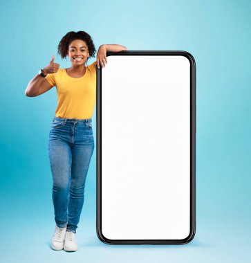 Mobil teklif. Mutlu siyah kadın boş beyaz ekranlı büyük bir cep telefonuna yaslanıyor, baş parmağını gösteriyor, yeni bir uygulama ya da web sitesi tavsiye ediyor, reklamınız için yer öneriyor,