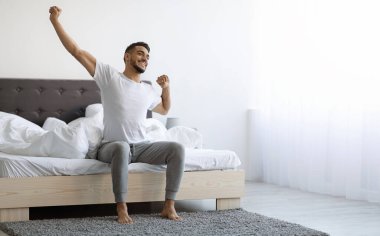 Good Morning. Happy Millennial Arab Guy Sitting On Bed And Stretching After Good Sleep, Handsome Smiling Young Middle Eastern Man Relaxing In Cozy Bedroom, Enjoying Home Comfort, Copy Space