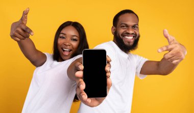 Yeni Mobil Uygulama Reklamı. Elinde boş ekranla akıllı telefonu tutan mutlu siyah çiftin portresi, model için boş alanı olan cihazları gösteriyor ve işaret ediyor, sarı stüdyo arka planı