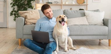 İçişleri Bakanlığı. Neşeli erkek serbest yazar oturma odasında dizüstü bilgisayar kullanarak oturuyor, köpeğini okşuyor, boşluğu kopyalıyor.