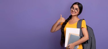 Mutlu Hintli kız öğrenci gözlük takıyor işaret işareti işareti yapıyor, kolej malzemeleri tutuyor ve sırt çantası takıyor, mor stüdyo arka planında tek başına duruyor bedava fotokopi alanı, afiş