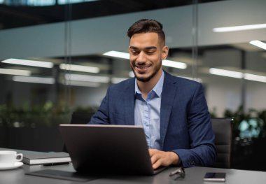 Gülümseyen genç ortadoğulu işadamı modern ofiste çalışıyor, bilgisayar klavyesine yazı yazıyor, iş ortaklarına e-posta gönderiyor, pazarlama araştırmaları yapıyor, uzay kopyaları kopyalıyor.