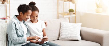 Evde kanepede telefon kullanan iki sevimli Afrikalı Amerikalı kız kardeş, akıllı telefon ekranına işaret ablası, boş alan