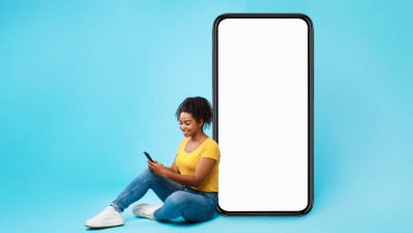Neşeli Afrikalı Amerikalı kadın boş ekranlı büyük bir cep telefonunun yanında oturuyor, mobil cihaz kullanıyor, mavi stüdyo arka planındaki yeni uygulamaları kontrol ediyor, web sitesi veya uygulama tasarımı için modelleme yapıyor.