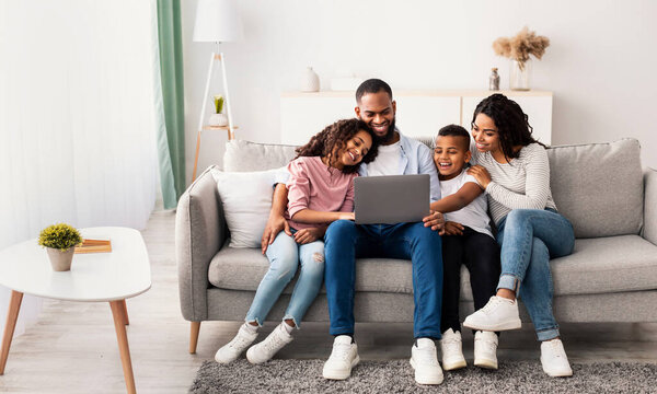 Family Weekend. Portrair of cheerful African American parents and their little children using laptop at home together, watching movie or browsing internet, sitting on the couch in living room