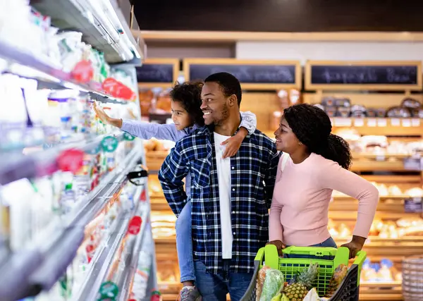 Marketten alışveriş yapan mutlu siyah aile portresi. Milenyum Afrikalı Amerikalı aileler ve güzel kızları büyük alışveriş merkezlerinde süt ürünlerini seçiyorlar.