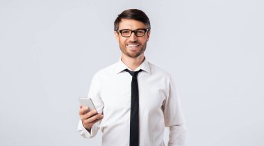 Beyaz gömlekli ve kravatlı bir adam akıllı telefon tutarken gülümsüyor. Beyaz bir arka planın önünde duruyor..