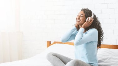 Neşeli genç Afrikalı kız yatak odasında kablosuz kulaklıkla müzik dinliyor, boş alanlı panorama.