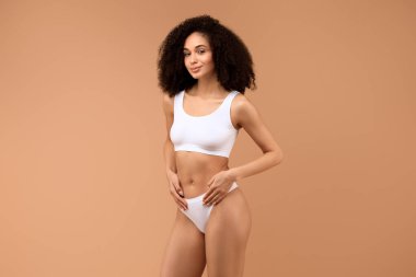 Beyaz sutyen ve iç çamaşırı giyen kıvırcık siyah saçlı genç bir kadın, bronz bir arka planın önünde duruyor, elleri kalçasında kameraya gülümsüyor..