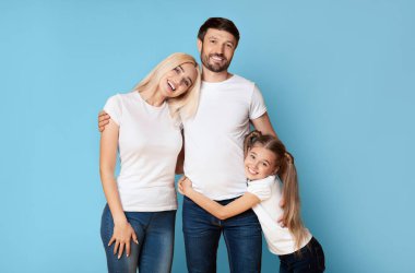 Üç kişilik gülümseyen bir aile, bir baba, bir anne ve küçük bir kız, mavi bir arkaplan önünde birlikte duruyorlar. Baba karısının belini tutarken, kız babasına sarılıyor.
