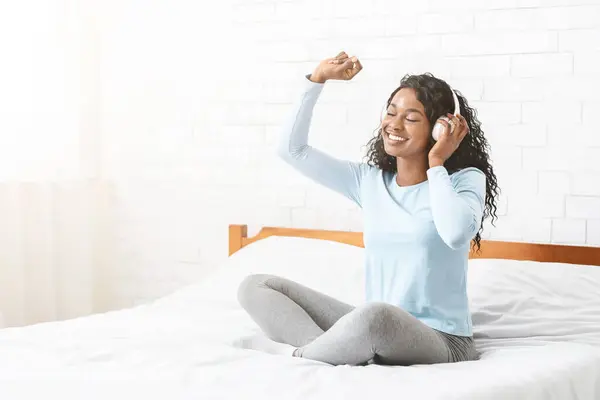 stock image Pretty black girl with closed eyes listening to music and dancing, sitting on bed, copy space